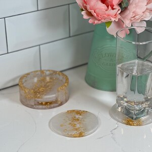 May include: Three white resin coasters with gold flecks. The coasters are round and have a raised edge. The coasters are on a white countertop.