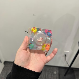 May include: A clear, square-shaped resin object filled with colorful miniature rubber ducks. A small, pink duck is encased in a clear, spherical bubble within the resin. A metal pin extends from one side of the object.