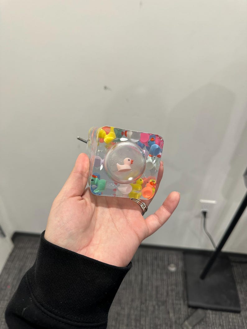 May include: A clear, square-shaped resin object filled with colorful miniature rubber ducks. A small, pink duck is encased in a clear, spherical bubble within the resin. A metal pin extends from one side of the object.