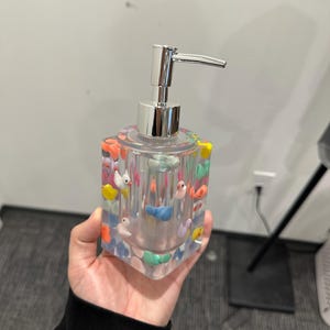 May include: Clear square soap dispenser with colorful duck figurines embedded in the resin. The dispenser has a shiny silver pump and is held in a hand. The ducks are various colors, including yellow, blue, and orange.
