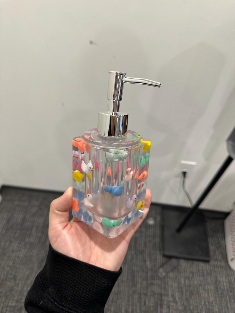May include: Clear square soap dispenser with colorful duck figurines embedded in the resin. The dispenser has a shiny silver pump and is held in a hand. The ducks are various colors, including yellow, blue, and orange.