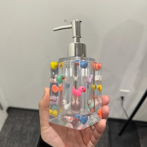 May include: Clear rectangular soap dispenser with a silver pump, decorated with colorful miniature ducks. The ducks are in various colors, including pink, yellow, blue, and orange. The dispenser is held in a hand.