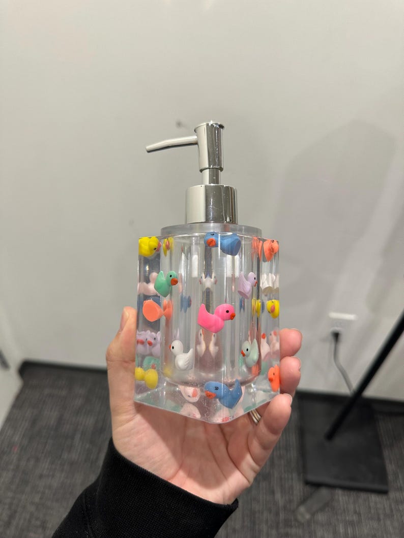 May include: Clear rectangular soap dispenser with a silver pump, decorated with colorful miniature ducks. The ducks are in various colors, including pink, yellow, blue, and orange. The dispenser is held in a hand.