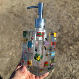 May include: Clear resin soap dispenser with a silver pump. The dispenser is decorated with miniature colorful gift boxes and a string of lights. The dispenser is held in a hand.