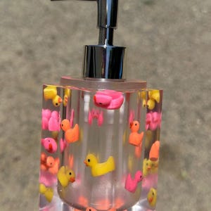 May include: A clear, rectangular soap dispenser with a chrome pump. The dispenser is filled with small, colorful rubber duckies in shades of yellow, pink, and orange. The base and top are pink.