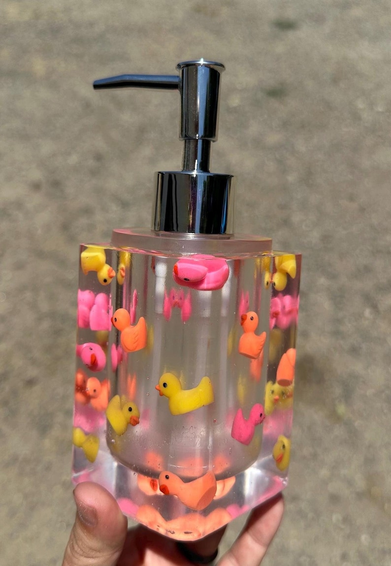 May include: A clear, rectangular soap dispenser with a chrome pump. The dispenser is filled with small, colorful rubber duckies in shades of yellow, pink, and orange. The base and top are pink.