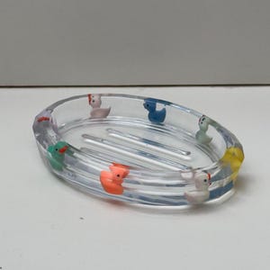 Puede incluir: Un jabonera ovalada transparente con un patrón de rejilla elevado en la parte inferior. La jabonera está decorada con patitos de goma de colores alrededor del borde.