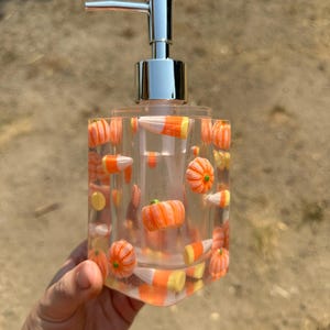 Pode incluir: Dispensador de sabonete quadrado transparente com uma bomba prateada. O dispensador é preenchido com doces de milho laranja e amarelo e pequenas abóboras laranja. Um item decorativo para o banheiro ou cozinha.