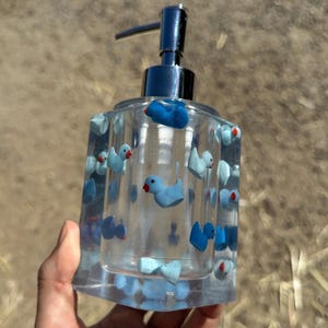 May include: Clear glass soap dispenser decorated with blue duck figurines and red accents. The dispenser has a silver pump and is held by a hand. The dispenser is a square shape.