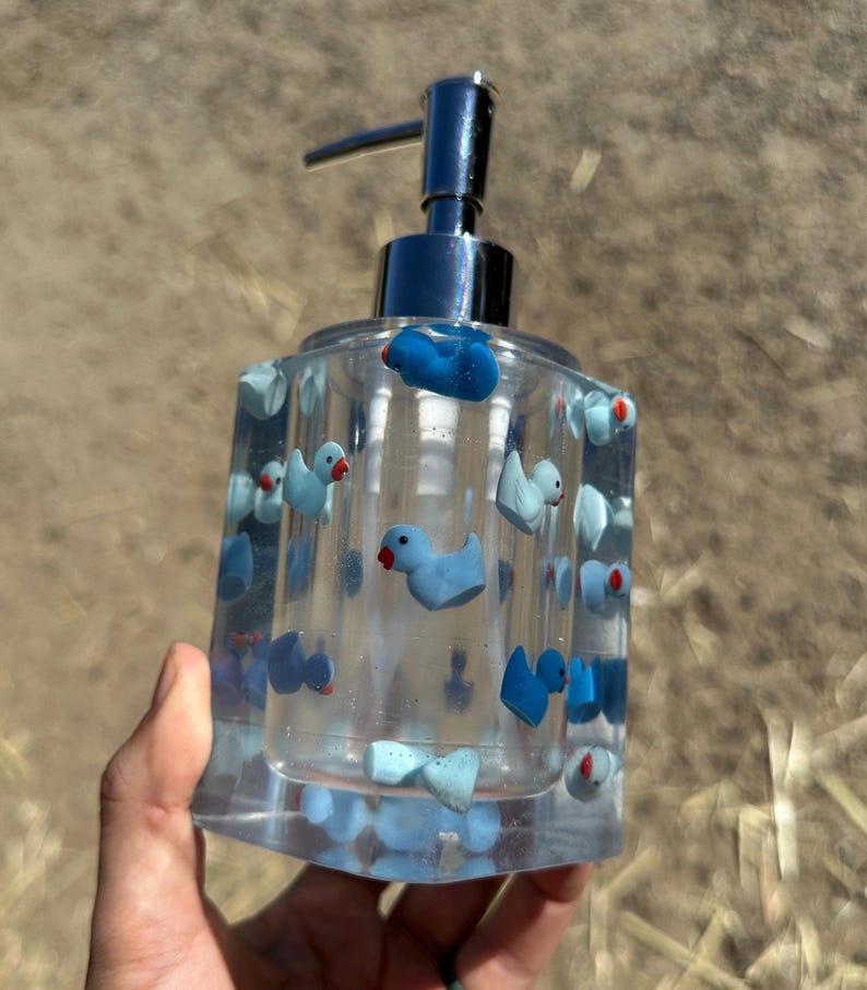 May include: Clear glass soap dispenser decorated with blue duck figurines and red accents. The dispenser has a silver pump and is held by a hand. The dispenser is a square shape.