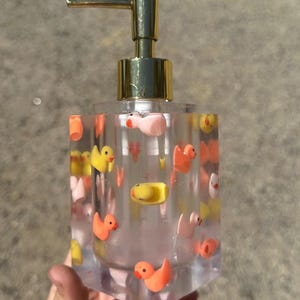 May include: Clear soap dispenser with a gold pump top, filled with small, colorful rubber duckies. The ducks are yellow, orange, and pink. The dispenser is held in a hand.