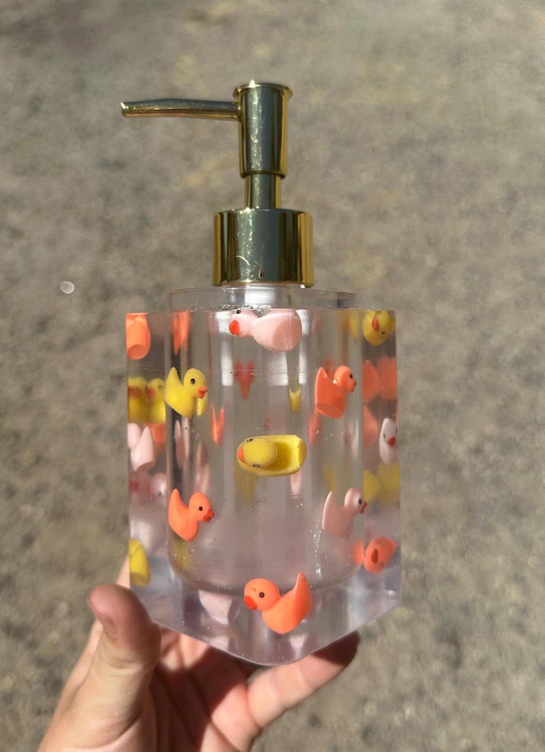 May include: Clear soap dispenser with a gold pump top, filled with small, colorful rubber duckies. The ducks are yellow, orange, and pink. The dispenser is held in a hand.