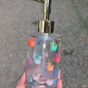 May include: Clear plastic soap dispenser with a gold-colored pump and a collection of colorful duck and flower decorations. The dispenser is filled with a clear liquid and is held in a hand.