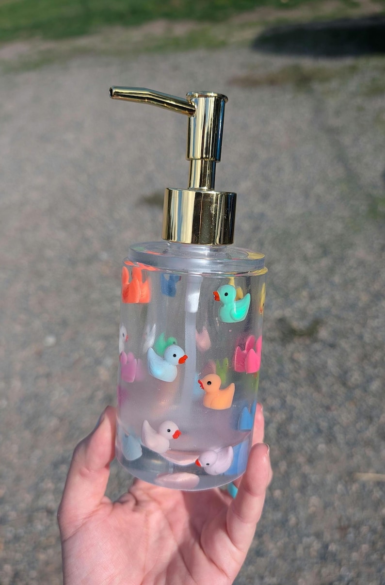 May include: Clear plastic soap dispenser with a gold-colored pump and a collection of colorful duck and flower decorations. The dispenser is filled with a clear liquid and is held in a hand.