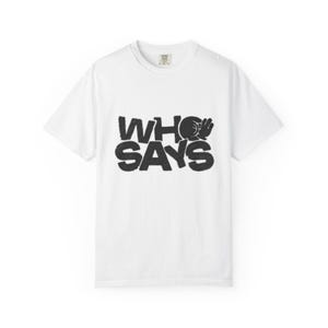 May include: White t-shirt with a crew neck and short sleeves. The shirt features the phrase "WHO SAYS" in a bold, black, distressed font. The "O" in "WHO" is replaced with a graphic of a hand making an "OK" gesture.
