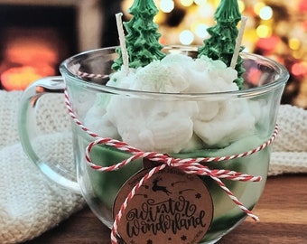 May include: A glass mug candle with a winter wonderland theme. The candle is decorated with green pine trees, white snow, and red and white twine. The candle has a brown label that says "winter wonderland" and "Sweet Candle House Co."