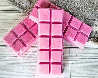 May include: Three pink wax melts in a rectangular shape with a grid pattern. The wax melts are on a white wooden surface.