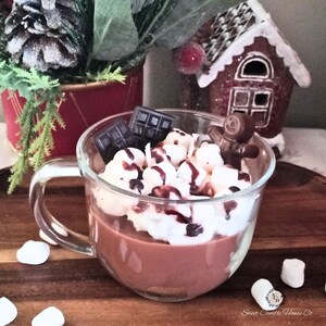 May include: A glass mug filled with a chocolate candle, topped with white frosting, chocolate squares, and a gingerbread man. The mug is sitting on a wooden surface with white marshmallows scattered around it.