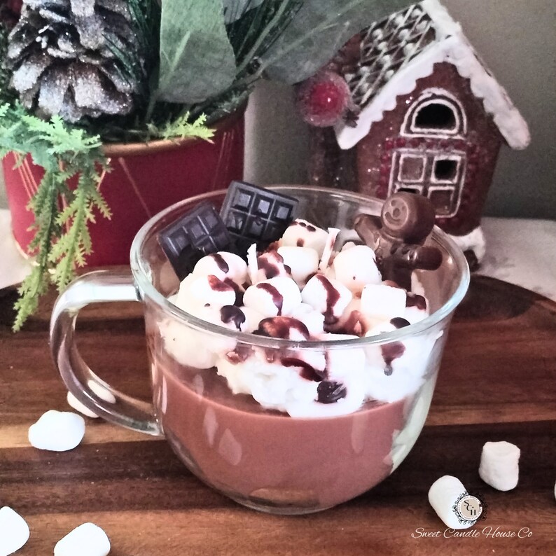 May include: A glass mug filled with a chocolate candle, topped with white frosting, chocolate squares, and a gingerbread man. The mug is sitting on a wooden surface with white marshmallows scattered around it.