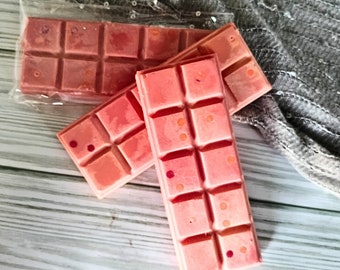 May include: Three pink wax melts in a clear plastic wrapper. The wax melts are divided into squares and have small orange and red flecks throughout.