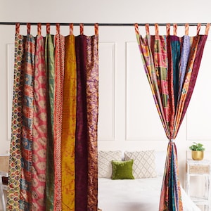 Peut inclure: Deux rideaux patchwork colorés en tissus différents sont suspendus à une tringle à rideaux noire. Les rideaux sont un mélange de tissus rouges, verts, jaunes, bleus et violets avec différents motifs. Les rideaux sont suspendus devant un mur blanc et un lit blanc avec un oreiller vert.