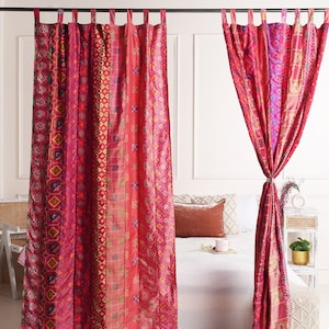May include: A pair of pink and red patterned curtains with a geometric design. The curtains are hung on a metal rod and are tied back on each side. The curtains are in front of a white bed with a white duvet and a red pillow.