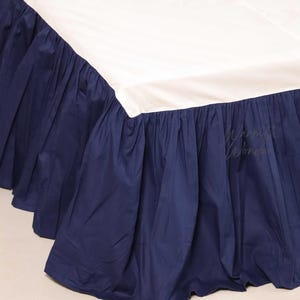 Może przedstawiać: Spódnica do łóżka z granatowym falbaną. Jest połączona z białym prześcieradłem. Granatowy materiał jest marszczony, tworząc teksturowany, warstwowy efekt. Ogólna estetyka jest klasyczna i elegancka.