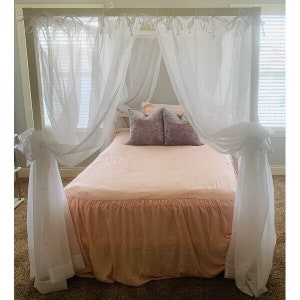 May include: A white canopy bed with a pink ruffled bed skirt and white sheer curtains. Two gray pillows are on the bed.