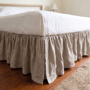 May include: A bed skirt in a natural beige color featuring a gathered, ruffled design that hangs from the base of the mattress. The fabric has a visible linen-like texture, and the skirt includes a vertical pleat detail along the side panel.