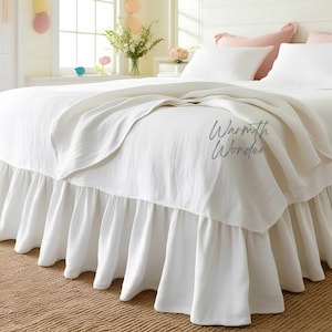 Puede incluir: Una cama blanca con una falda de cama con volantes y una colcha blanca a juego. La cama está hecha con almohadas blancas y una manta doblada. El texto "Warmth Wonder" es visible en la manta.