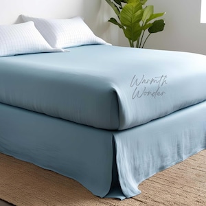 Könnte beinhalten: Ein hellblaues Bettwäsche-Set mit Spannbettlaken, Bettrock und zwei weißen Kissen mit Muster. Der Text "Warmth Wonder" ist auf das Spannbettlaken gedruckt. Das Bett steht auf einem gewebten Teppich.