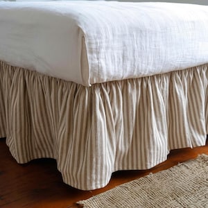 Puede incluir: Un faldón de cama con un diseño fruncido y con volantes con rayas verticales de color beige y blanco. El faldón se coloca debajo de una colcha blanca lisa y texturizada que cae sobre el borde superior del marco de la cama, creando un aspecto en capas.