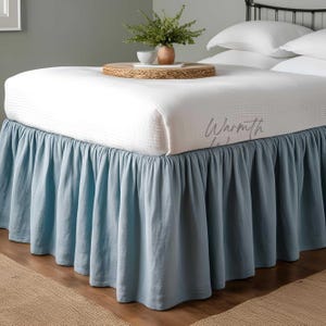 May include: A light blue bed skirt with a ruffled edge. The bed skirt is made of a soft, linen-like fabric. The bed skirt is shown on a white bed with a white headboard.