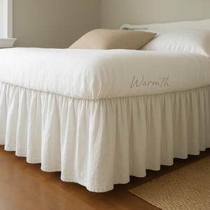 May include: A white bed skirt with a ruffled design. The bed is covered with a white comforter and pillows. The word "Warmth" is written on the comforter in a cursive font. The bed is in a bedroom with a neutral color scheme.