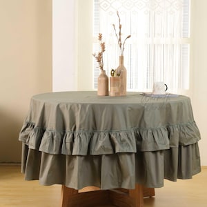 May include: A round, sage green tablecloth with a ruffled skirt. The table is set with decorative vases and a mug. The text "Warmth Wonder" is visible on the table. The table is made of wood.