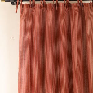 May include: A red and white striped curtain panel with tie-top details. The curtain hangs from a black rod with gold accents. The fabric features vertical stripes, creating a classic and simple design. The curtain is made of a soft, textured material.