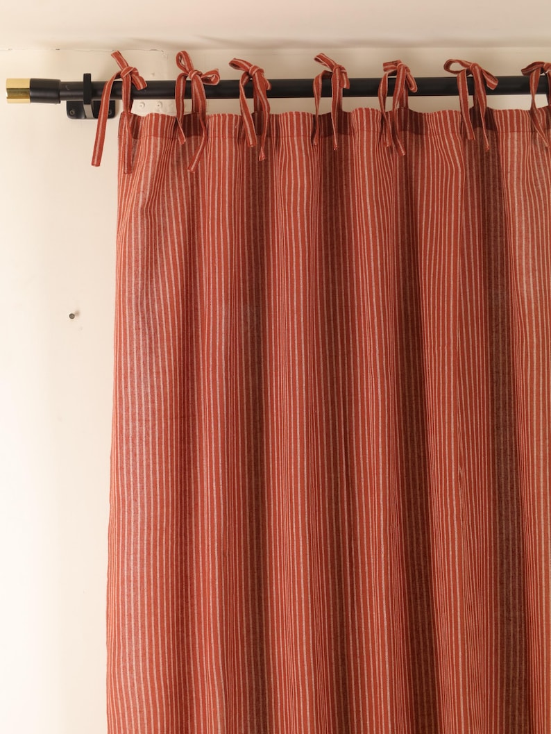 May include: A red and white striped curtain panel with tie-top details. The curtain hangs from a black rod with gold accents. The fabric features vertical stripes, creating a classic and simple design. The curtain is made of a soft, textured material.