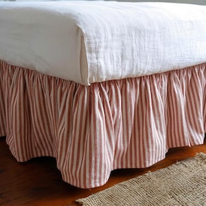 May include: A bed valance featuring a vertical striped pattern in muted red and white. The fabric is gathered to create a ruffled effect along the base of the bed, which is covered by a plain, off-white top sheet with a visible woven texture.
