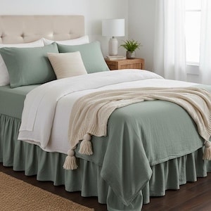May include: A bed with sage green bedding, including a fitted sheet, duvet cover, and ruffled bed skirt. The bed is made with pillows and a cream-colored throw blanket with tassels. A beige upholstered headboard completes the look.