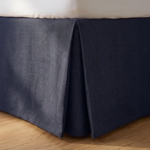 May include: A dark navy blue bed skirt with a textured fabric finish and a pleated corner design. The bed skirt is positioned beneath a white mattress, featuring a clean, straight-lined structure that covers the base of the bed frame.