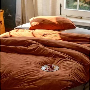 以下が含まれることがあります： 温かいさび色の羽毛布団カバーに白い枕と、赤いザクロの種が載った小さな白い皿が置かれています。寝具は白いシーツと白いレースのトリムが付いたベッドの上に置かれています。テキスト「Warmth Wonder」は白地に黒で書かれています。