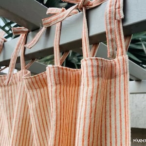 Puede incluir: Tres cortinas de tela a rayas naranja y blanco colgadas de una pérgola de madera. Las cortinas tienen lazos en la parte superior.
