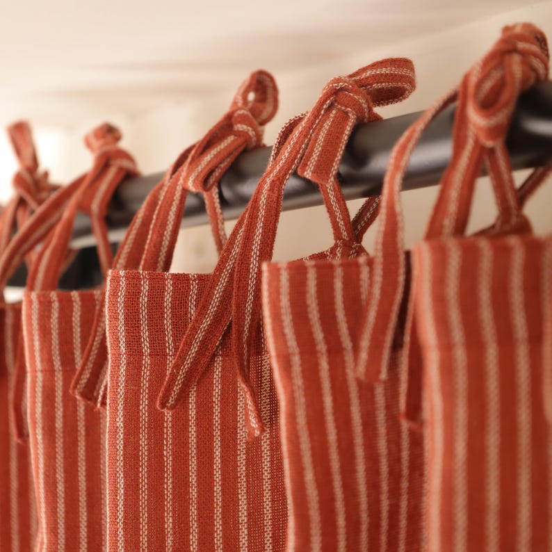 May include: Close-up of several terracotta-colored curtains with white vertical stripes. The curtains are hanging from a black rod and tied with matching fabric ties. The fabric has a textured appearance, suggesting a linen or similar material.