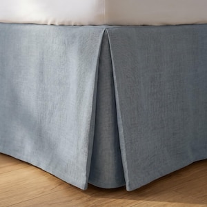May include: A dusty blue bed valance with a textured fabric finish, featuring a tailored design with a single box pleat at the corner. The valance hangs from the base of a bed, providing a clean, structured look that covers the lower frame of the bed.