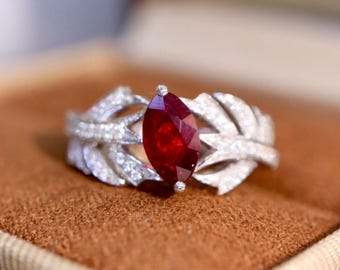 May include: A silver-toned ring featuring a central marquise-cut deep red gemstone. The band is designed with a textured, leaf-like motif encrusted with numerous small, clear pavé-set stones that extend along both sides of the central setting.