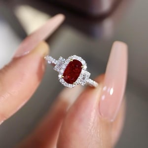 Könnte beinhalten: Diamantring mit einem großen, oval geschliffenen, tiefroten Edelstein als Mittelpunkt. Der Edelstein ist von einem Kranz kleiner, runder Diamanten umgeben, mit zusätzlichen Diamantakzenten auf dem Band. Der Ring ist aus weißem Metall.