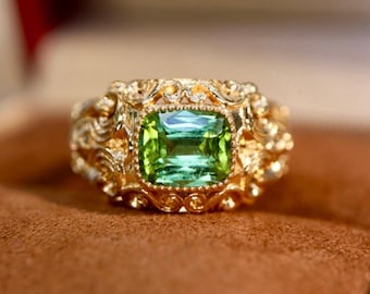 Può includere: Un anello d'oro dal design ornato, con una grande pietra preziosa verde chiaro a taglio quadrato. La fascia e l'incastonatura dell'anello sono finemente dettagliate con motivi a spirale. La gemma ha una leggera sfumatura blu.