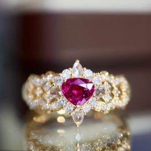 May include: A gold ring featuring a heart-shaped, deep pink gemstone at its center, surrounded by smaller, clear gemstones. The band is intricately designed with additional clear stones.