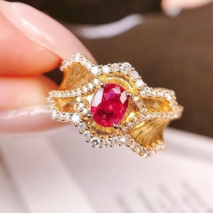 May include: A gold ring with a large oval ruby gemstone surrounded by smaller diamonds. The ring has a detailed design with a wavy band.
