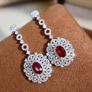 Puede incluir: Un par de pendientes colgantes. Cada pendiente presenta una gran gema roja oscura de corte ovalado en el centro, rodeada por un halo de pequeñas gemas blancas brillantes. Los pendientes tienen un diseño floral y están engastados en metal plateado.